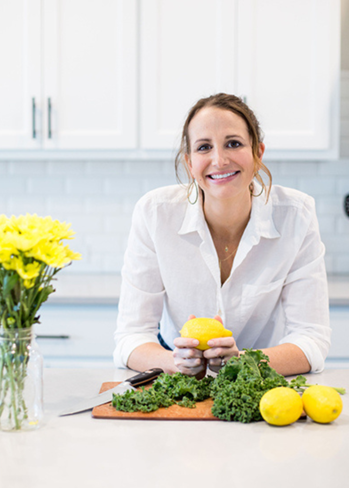 Lacey Baier, nutrition and fitness coach and founder of Cleanish, smiling in her kitchen while holding fresh lemons beside leafy greens, representing her practical, real-life approach to healthy cooking and meal prep.